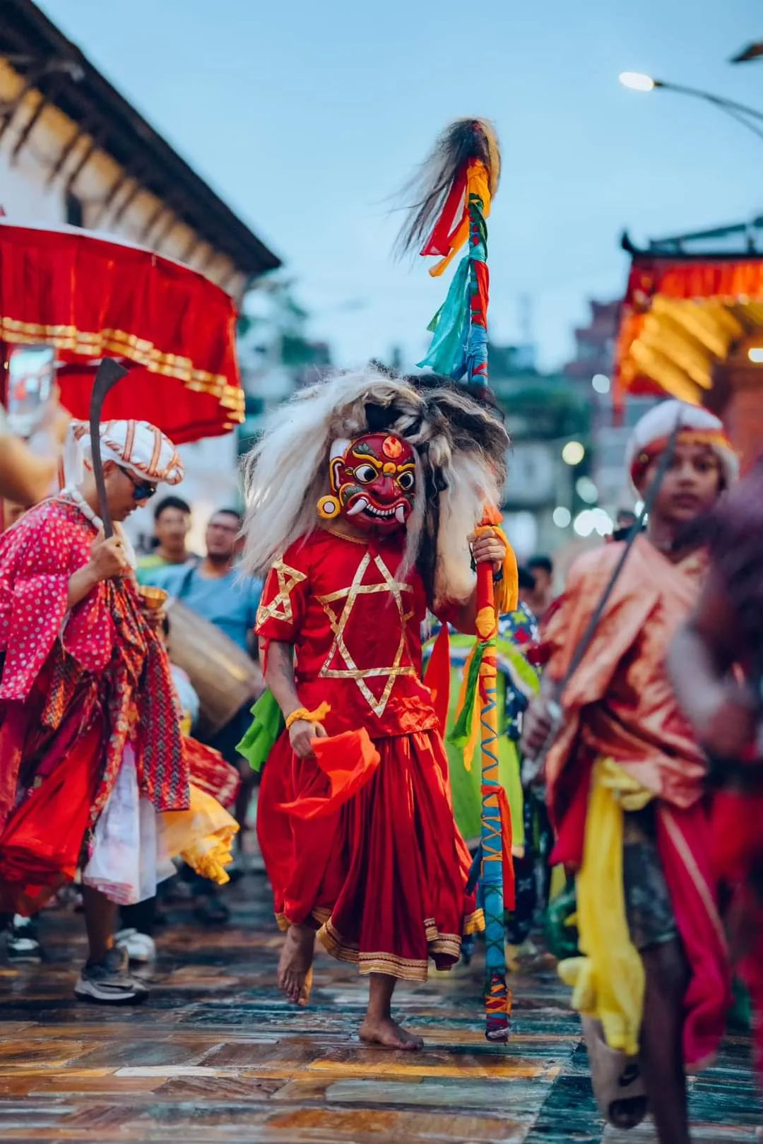 A lively procession of people in bright costumes, featuring a striking red lakhey, strolls along the street.